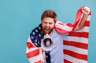 Heyecanlı, kendine güvenen sakallı bir adamın, Amerika Birleşik Devletleri bayrağını taşıyan hoparlöre bağırışının portresi, vatansever protesto. Mavi arka planda kapalı stüdyo çekimi.