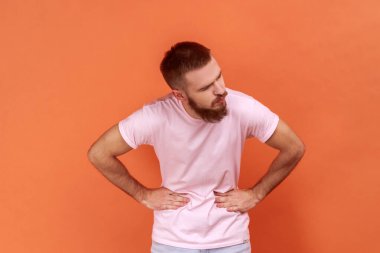 Sağlıksız sakallı hasta bir adamın portresi. Karın ağrısı, şiddetli karın ağrısı, pembe tişört giymesi. Turuncu arka planda kapalı stüdyo çekimleri.