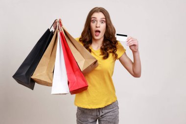 Alışveriş kredisi ve nakit para! Sarı tişörtlü, şok olmuş kıvırcık saçlı genç kız portresi elinde kredi kartı ve paketler, ağzını açık tutuyor. Gri arka planda izole edilmiş kapalı stüdyo görüntüleri.