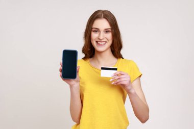 Sarı günlük tişörtlü neşeli bir genç kız. Elinde boş akıllı telefon ve kredi kartı var. Cep telefonu bankacılığından memnun. Gri arka planda izole edilmiş kapalı stüdyo görüntüleri.
