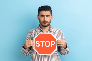 Ciddi sakallı iş adamının portresi yasağın sembolü olarak trafik işaretini tutuyor, kısıtlamalar, kameraya bakıyor, çizgili gömlek giyiyor. Mavi arka planda kapalı stüdyo çekimi.