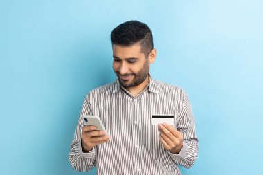 Akıllı telefon ve kredi kartı tutan mutlu sakallı iş adamının portresi, veri girişi, online alışveriş, çizgili gömlek. Mavi arka planda kapalı stüdyo çekimi.