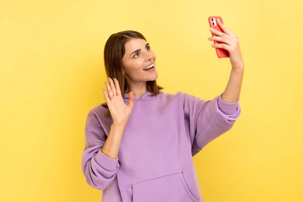 一个积极漂亮的女人对着智能手机挥手和自拍的画像 一边打电话一边给她打手势 穿着紫色的帽衫 在黄色背景下隔离的室内拍摄 — 图库照片