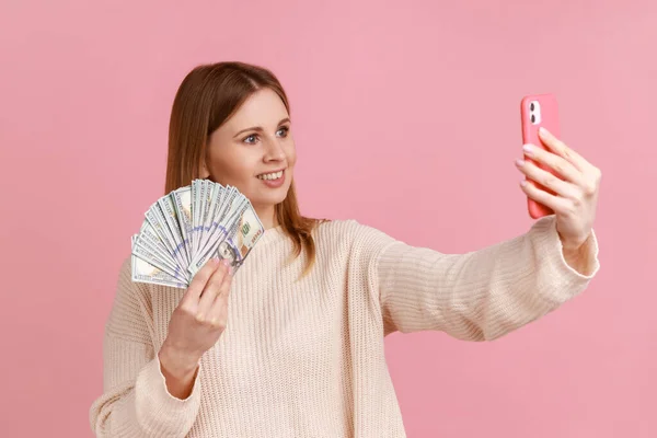 彼女の給料や宝くじの勝利を誇り 白いセーターを着て ドル札で自撮りを取って肯定的な豊かな愛らしいブロンドの女性の肖像画 ピンクの背景に隔離された屋内スタジオショット — ストック写真