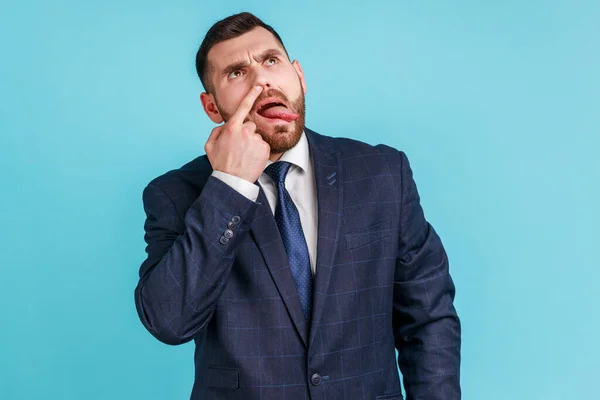 Misconduct, bad manners. Funny man wearing official style suit picking nose and sticking out tongue with dumb comical expression, fooling around. Indoor studio shot isolated on blue background.