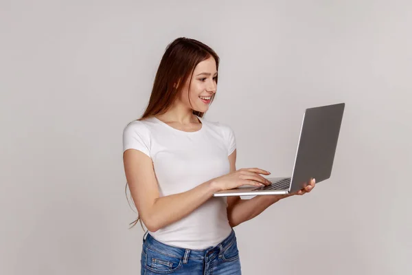 ノートパソコンを持っているかなり肯定的な女性 キーボードを入力します サーフィンインターネット ノートブックを使用して作業学生やフリーランス 白いTシャツを着て 灰色の背景に隔離された屋内スタジオショット — ストック写真