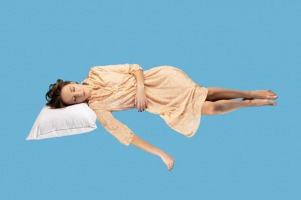 Sleeping beauty hovering in air. Relaxed girl in yellow dress lying comfortably on pillow levitating, keeping eyes closed, watching dreams. full length studio shot isolated on blue background, indoor