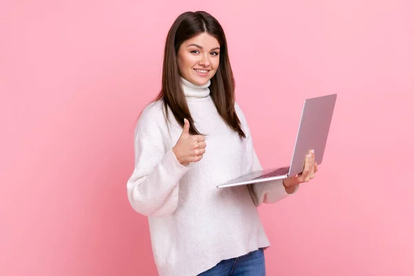 幸せな女性のフリーランサーは 白いカジュアルなスタイルのセーターを着て 彼女の仕事が好き 親指を表示し カメラを見て ラップトップコンピュータ上で動作します ピンクの背景に隔離された屋内スタジオショット — ストック写真