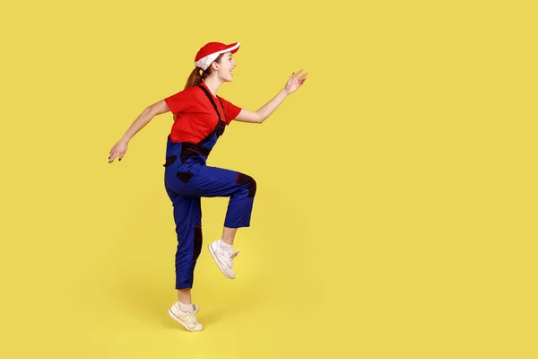 前向きな労働者の女性の側のビューを1つの足の上に腕を上げて立って 先を見て 行進し 幸せな表情は 全体と赤のキャップを着用 黄色の背景に隔離された屋内スタジオショット — ストック写真