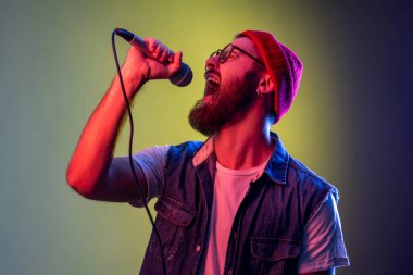Sakallı hippi yüksek sesle mikrofonu elinde tutarak şarkı söylüyor, karaoke yaparken dinleniyor, şarkıcı performansı sergiliyor, bere takıyor. Renkli neon ışıklı arka planda kapalı stüdyo çekimleri