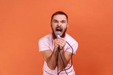 Elinde mikrofonla kameraya bakıp yüksek sesle bağıran pembe tişört giyen kızgın, sakallı adamın portresi. Turuncu arka planda kapalı stüdyo çekimleri.