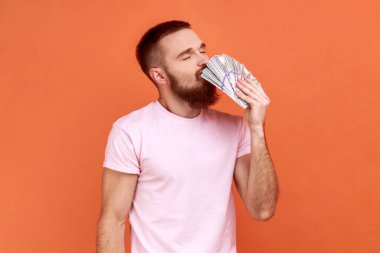 Sakallı adamın portresi dolar banknotları kokan, zevk ifadeli, büyük paralar kazanan, piyango kazanan, pembe tişört giyen. Turuncu arka planda kapalı stüdyo çekimleri