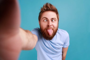 Yakışıklı çocuksu sakallı blog yazarının portresi. Bakış açısını değiştiriyor, dil gösteriyor, çocukça davranışlar sergiliyor, POV. Mavi arka planda kapalı stüdyo çekimi.