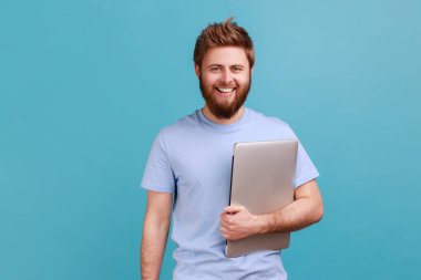 Bilgisayarı kapalı tutan sakallı adamın portresi ve dişlek gülüşüyle kameraya bakıp işi bitirmekten mutlu olmak. Mavi arka planda kapalı stüdyo çekimi.