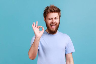 Sakallı adamın portresi tamam işareti, başarı işareti, iyi hizmetten memnun, kameraya bakıyor ve göz kırpıyor, pozitif duyguları ifade ediyor. Mavi arka planda kapalı stüdyo çekimi.