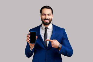 Sakallı adam akıllı telefonu boş ekranla işaret ediyor, kameraya gülümsüyor, reklam için boş alana bakıyor, resmi tarz takım elbise giyiyor. Gri arka planda izole edilmiş kapalı stüdyo görüntüleri.