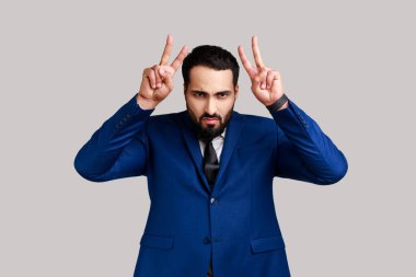 Çılgın sakallı adam boğa boynuzu hareketi yapıyor, parmakları başının üstünde, saldırgan ve tehditkar görünüyor, resmi stil kıyafeti giyiyor. Gri arka planda izole edilmiş kapalı stüdyo görüntüleri.