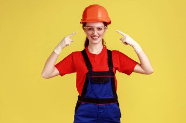 Koruyucu gözlükleri ve kaskı işaret eden, kameraya bakan, tulum giyen, gülümseyen olumlu müteahhit kadın portresi. Sarı arka planda kapalı stüdyo çekimleri.