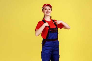 Gülümseyen, becerikli bir kadının portresi kendini gururlu bir yüz ifadesiyle gösteriyor başarısıyla övünüyor, tulum giyiyor ve kırmızı şapka takıyor. Sarı arka planda kapalı stüdyo çekimleri.