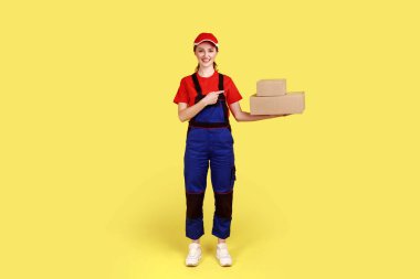 Ellerindeki karton paketleri gösteren güler yüzlü kurye kadın portresi, hızlı teslimat servisi, tulum ve kırmızı şapka giyiyor. Sarı arka planda kapalı stüdyo çekimleri.