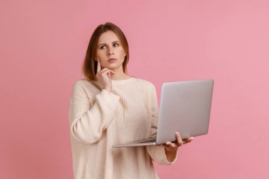 Düşünceli, sarışın bir kadının portresi dizüstü bilgisayarda çalışıyor, başka tarafa bakıyor, önemli bir projeyi düşünüyor, beyaz kazak giyiyor. Pembe arka planda kapalı stüdyo çekimleri.