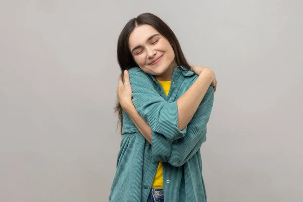 Positive self-esteem. Woman embracing herself and smiling with expression of pleasure, being selfish and narcissistic, wearing casual style jacket. Indoor studio shot isolated on gray background.