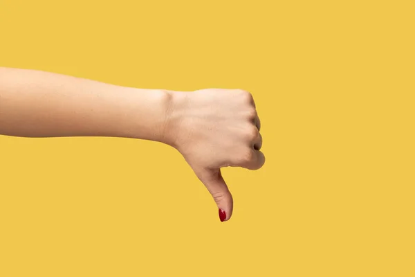 Closeup woman hand showing thumb down, rejection symbol, negative feedback. Indoor studio shot isolated on yellow background.