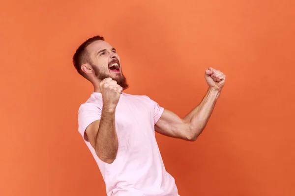 大胡子英俊男子的画像 他满脸喜色 挥动拳头和尖叫 庆祝胜利 身穿粉色T恤 表现出胜利的姿态 在橙色背景下被隔离的室内拍摄 — 图库照片