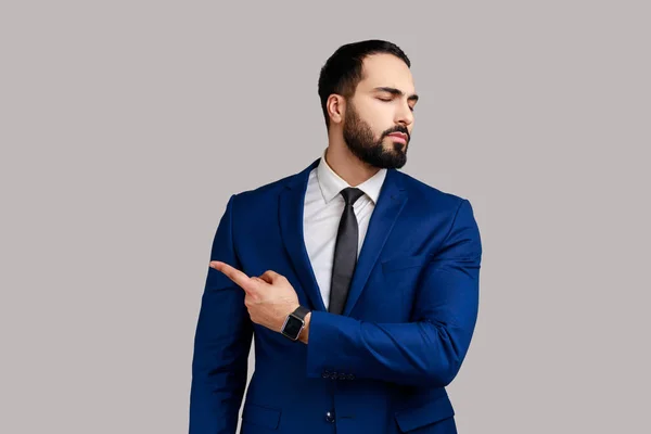 Portrait of bearded man pointing to the side and turning away with angry vexed face, giving order to leave, wearing official style suit. Indoor studio shot isolated on gray background.