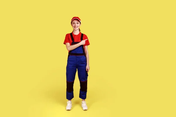 Full length portrait of optimistic handy woman standing and pointing finger away, showing copy space for advertisement, wearing overalls and red cap. Indoor studio shot isolated on yellow background.