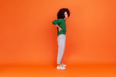 Afro saçlı, yeşil bir süveter giyen, sırtı ağrıyan, böbrekleriyle sorunu olan bir kadının uzun bir portresi. Turuncu arka planda kapalı stüdyo çekimleri.