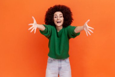 Afro saç stili, yeşil bir süveter giyen, elini kameraya uzatan mutlu bir kadının portresi kucaklaşacak. Turuncu arka planda kapalı stüdyo çekimleri.