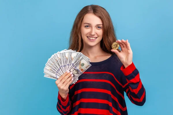 縞模様のカジュアルなスタイルのセーターを着た女性を喜ばせ 紙幣と金のビットコインのファンを示し 暗号通貨を紙幣と比較してください 青い背景に隔離された屋内スタジオショット — ストック写真