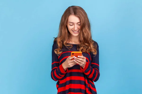 幸せな女性は 携帯電話を使用して快適な笑顔で縞模様のカジュアルなスタイルのセーターを着て ソーシャルネットワークでのメッセージに答える 記事を書く 青い背景に隔離された屋内スタジオショット — ストック写真