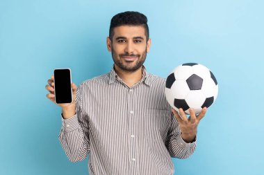 Futbol topu tutan sakallı, mutlu bir adam ve akıllı telefon boş siyah ekran, şampiyonluk için bilet rezervasyonu, çizgili gömlek giyiyor. Mavi arka planda kapalı stüdyo çekimi.