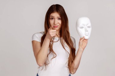 Portrait of dark haired woman pointing finger on you, holding white mask with unknown face, blaming you in duplicity, wearing white T-shirt. Indoor studio shot isolated on gray background. clipart