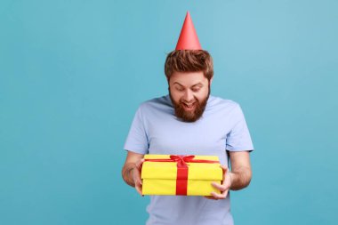 Parti konisinde elinde sarı paketlenmiş hediye kutusu olan heyecanlı sakallı adamın portresi tatmin olmuş bir ifadeyle şu ana bakıyor. Mavi arka planda kapalı stüdyo çekimi.