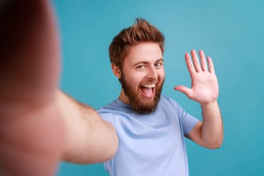 Portrait of positive friendly bearded man blogger making selfie or streaming, waving hand POV, point of view of photo, saying hello or bye. Indoor studio shot isolated on blue background.