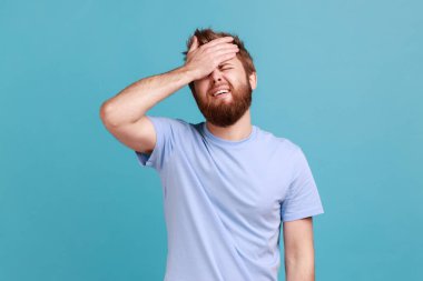 Üzgün sakallı adamın portresi elinde fotojenik bir jestle duruyor, kendini suçluyor, kötü anılar yüzünden üzüntü duyuyor. Mavi arka planda kapalı stüdyo çekimi.