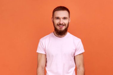 Kameraya bakıp gülümseyen mutlu sakallı adamın portresi, olumlu duygular ifade eden, iyi ruh halinde olan, pembe tişört giyen. Turuncu arka planda kapalı stüdyo çekimleri.