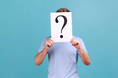Portrait of unknown bearded man hiding his face behind white paper with question mark, finding smart solution, asking for advice. Indoor studio shot isolated on blue background.