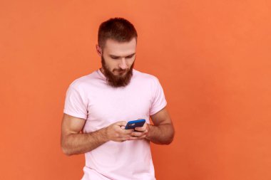 Sakallı bir adamın portresi, sakin bir ifadeyle cep telefonu kullanıyor, akıllı telefona bağımlı, sosyal ağda mesajlaşıyor, pembe tişört giyiyor. Turuncu arka planda kapalı stüdyo çekimleri.