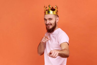 Portrait of bearded man looking smiling at camera, posing with crown on head, pointing at you with happy expression, wearing pink T-shirt. Indoor studio shot isolated on orange background.
