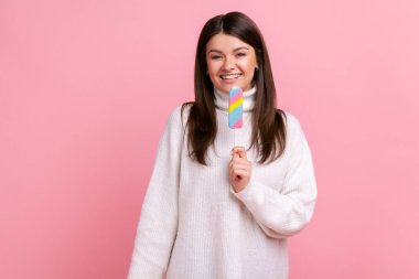 Tatmin olmuş gülümseyen esmer kadın elinde lezzetli dondurmayla kameraya mutlu bir ifadeyle bakıyor, beyaz bir süveter giyiyor. Pembe arka planda kapalı stüdyo çekimleri.