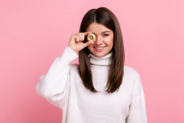 İyimser, pozitif esmer kadın, gözünü altın bitcoin ile kaplıyor, kameraya gülümsüyor, beyaz, sıradan bir süveter giyiyor. Pembe arka planda kapalı stüdyo çekimleri.
