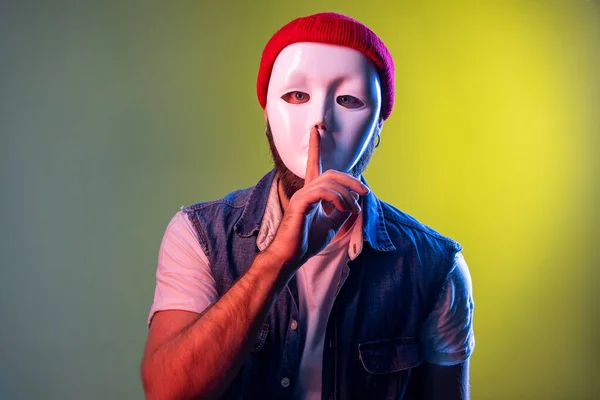 Anonymous hipster man in white mask on face, hiding personality, conspiracy and privacy, keeping finger near lips, secrets. Indoor studio shot isolated on colorful neon light background.