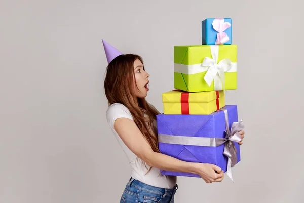 Portrait of excited woman holding stack of presents, having surprised excited facial expression, keeps mouth opened, wearing white T-shirt. Indoor studio shot isolated on gray background.
