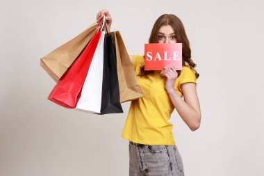 Portrait of beautiful brown haired teenager girl in yellow T-shirt holding shopping bags and covering half of face with card with Sale inscription. Indoor studio shot isolated on gray background.