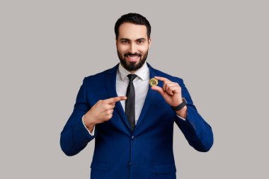 Memnuniyet duyan sakallı iş adamının portresi altın Bitcoin 'i işaret ediyor, ekommerce, resmi stil takım elbise giyiyor. Gri arka planda izole edilmiş kapalı stüdyo görüntüleri.
