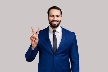 Sakallı adam barışın sembolünü parmaklarıyla gösteriyor, dişlek bir gülümsemeyle kameraya bakıyor, başarılı zaferini kutluyor, takım elbise giyiyor. Gri arka planda izole edilmiş kapalı stüdyo görüntüleri.